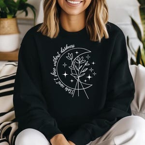 May include: Black sweatshirt with a white graphic of a crescent moon, flowers, and stars. The text "Stars can't shine without darkness" is written around the moon. The sweatshirt is a crew neck style.