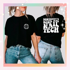 May include: Black t-shirt with the text "SOMEBODY'S BOMB ASS NAIL TECH" in white on the back. The front of the shirt has a smiley face design with the words "BOMB ASS NAIL TECH" in a circle. The model is wearing blue jeans.
