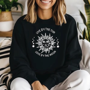 May include: Black sweatshirt featuring a white sun and moon design. The sun has a smiling face, surrounded by rays and stars. The text "LIVE BY THE SUN LOVE BY THE MOON" is printed above and below the sun.