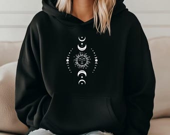 May include: Black hooded sweatshirt with a white celestial design. The design features a sun with a face, surrounded by phases of the moon and small dots. The sweatshirt has a front pocket and a drawstring hood.