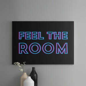May include: A rectangular black wall art piece featuring the text FEEL THE ROOM in a bold, blocky font. The letters have a layered, three-dimensional effect with bright cyan and magenta outlines against the solid black background.