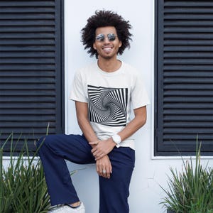May include: A short-sleeved, off-white crew neck t-shirt featuring a large, centered black and white graphic print of a swirling, hypnotic vortex pattern. The shirt is paired with solid navy blue trousers and a white wristwatch on the wrist.