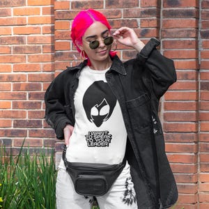 May include: A white t-shirt featuring a black graphic of an alien head silhouette above the text I WANT TO SPEAK TO YOUR LEADER. The shirt is worn under a black button-down shirt and paired with a black fanny pack, gold hoop earrings, and sunglasses.