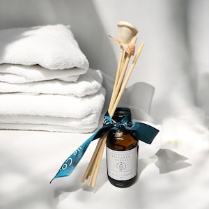 May include: A brown glass bottle with a teal and white label that says "Diffuser Refill" and "Fire & Grace Candle Co.". The bottle is surrounded by wooden sticks and a teal ribbon. The bottle is sitting on a white surface.