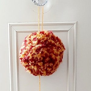 May include: A handmade yarn wall hanging with three decorative elements. The top is a round, multi-coloured pom-pom. Below is a yellow tassel, and at the bottom is a smaller, two-toned tassel. The hanging is suspended from a crystal knob.