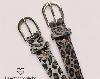 May include: Two leopard print belts with gold-coloured buckles. The belts have a brown and black spotted pattern. The belts are displayed on a light pink background. The text "Handtaschenliebe" is at the bottom left.