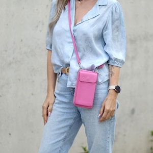 May include: A bright pink leather crossbody phone bag with a matching strap. The bag has a flap closure and is worn across the body. The bag is paired with a light blue button-down shirt and light wash jeans.