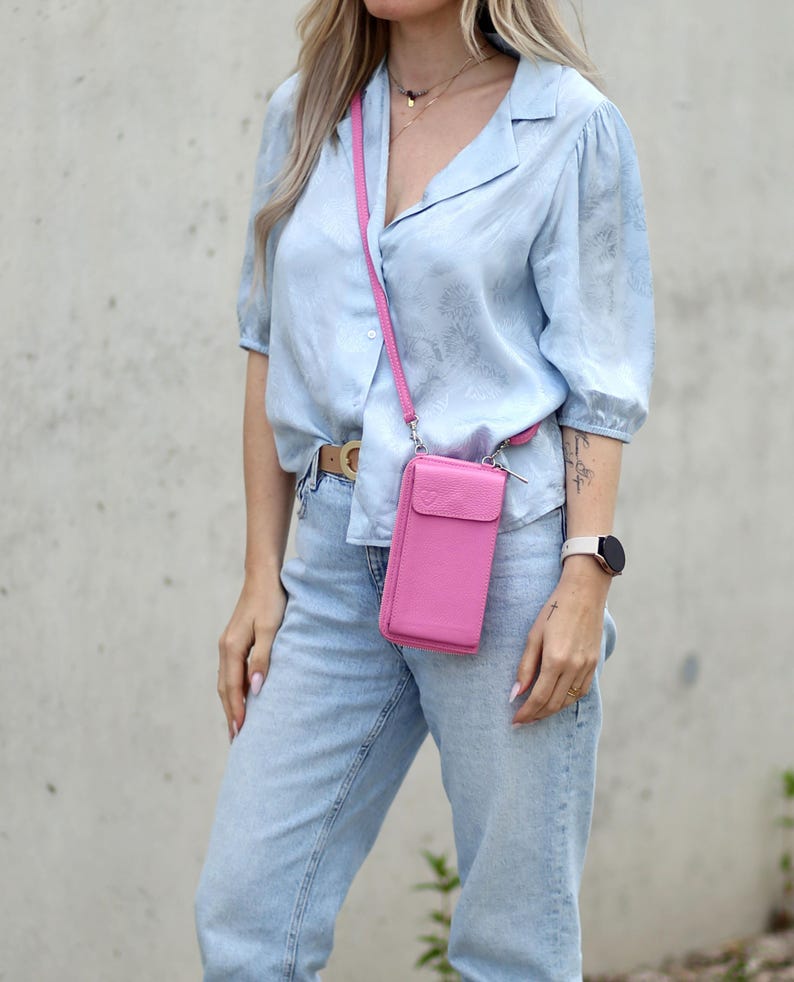 May include: A bright pink leather crossbody phone bag with a matching strap. The bag has a flap closure and is worn across the body. The bag is paired with a light blue button-down shirt and light wash jeans.