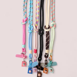 Könnte beinhalten: Eine Sammlung farbenfroher Handy-Lanyards. Jedes Lanyard hat ein anderes Farbschema, darunter Pink, Blau, Schwarz und gemusterte Optionen. Die Lanyards haben ein Knotendesign und sind mit kleinen, dekorativen Anhängern verziert.