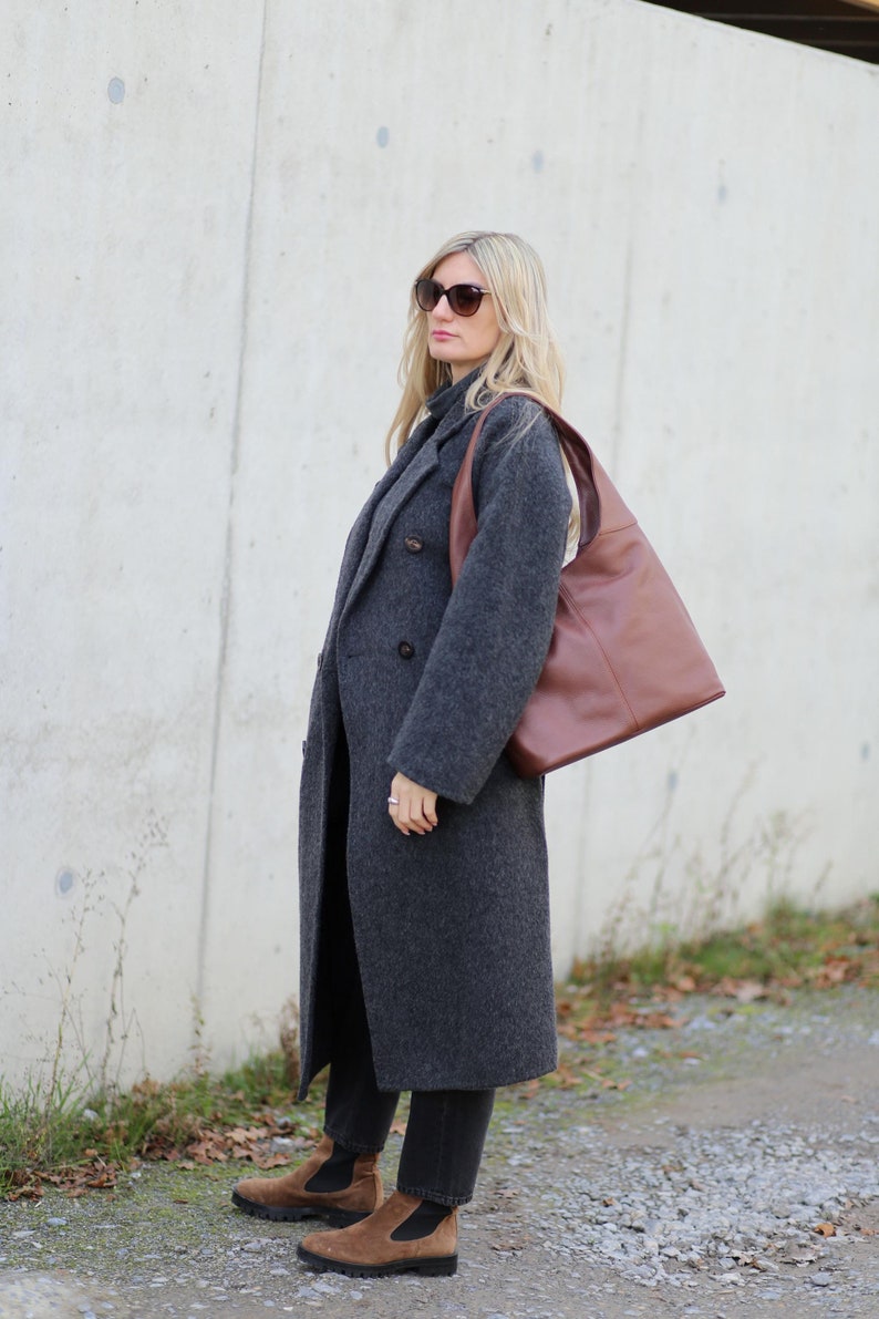 May include: A woman wearing a grey wool coat, black jeans, brown suede ankle boots, and sunglasses. She is carrying a brown leather tote bag.