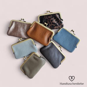 Può includere: Un assortimento di piccoli portamonete in pelle di vari colori, tra cui marrone, blu e nero. Ogni portamonete ha una chiusura in metallo e una superficie strutturata. I portamonete sono disposti su uno sfondo rosa chiaro.