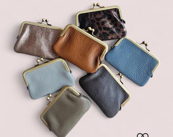 May include: An assortment of small, leather coin purses in various colors, including brown, blue, and black. Each purse has a metal clasp and a textured surface. The purses are arranged on a light pink background.