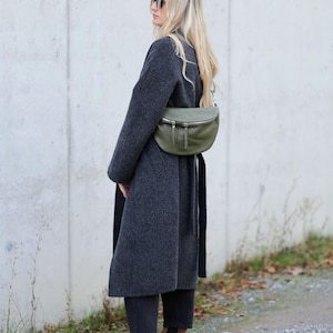 Op de afbeelding: Een persoon draagt een lange, donkergrijze jas en een zonnebril, met een groene leren heuptas. Ze draagt ook een zwarte broek en bruine su&egrave;de Chelsea boots. De achtergrond is een lichtgrijze muur en een grindpad.