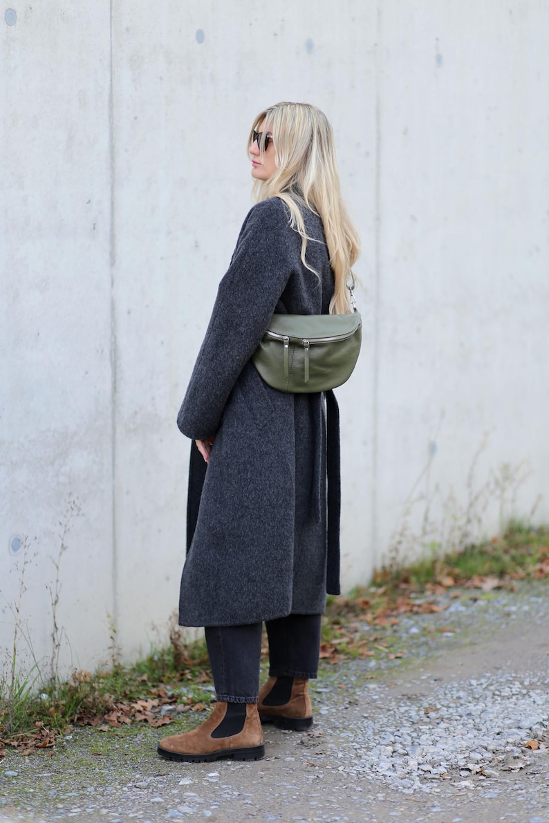 Op de afbeelding: Een persoon draagt een lange, donkergrijze jas en een zonnebril, met een groene leren heuptas. Ze draagt ook een zwarte broek en bruine su&egrave;de Chelsea boots. De achtergrond is een lichtgrijze muur en een grindpad.