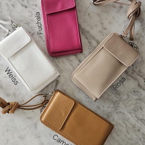 May include: Four leather crossbody phone cases in white, fuchsia, tan, and camel. Each case has a flap closure, a zipper, and a detachable strap. The names "Weiss", "Fuchsia", "Rosa", and "Camel" are printed on the cases.