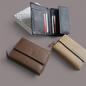 Puede incluir: Una variedad de carteras de cuero en negro, marrón y dorado. La cartera negra está abierta, revelando ranuras para tarjetas. Una caja de regalo con un patrón de confeti colorido está en el fondo. Las carteras tienen un acabado texturizado.