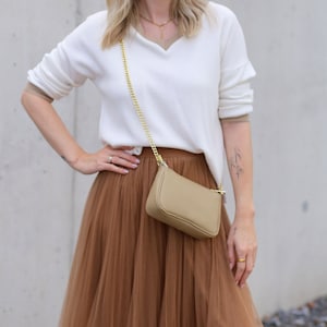 May include: A woman wearing a white sweater with a brown tulle skirt and a small beige crossbody bag with a gold chain strap.