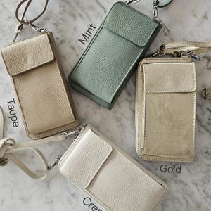 May include: Four leather crossbody phone bags in taupe, mint, gold, and creme. Each bag has a flap closure, a zipper, and a long strap. The bags are displayed on a marble surface.