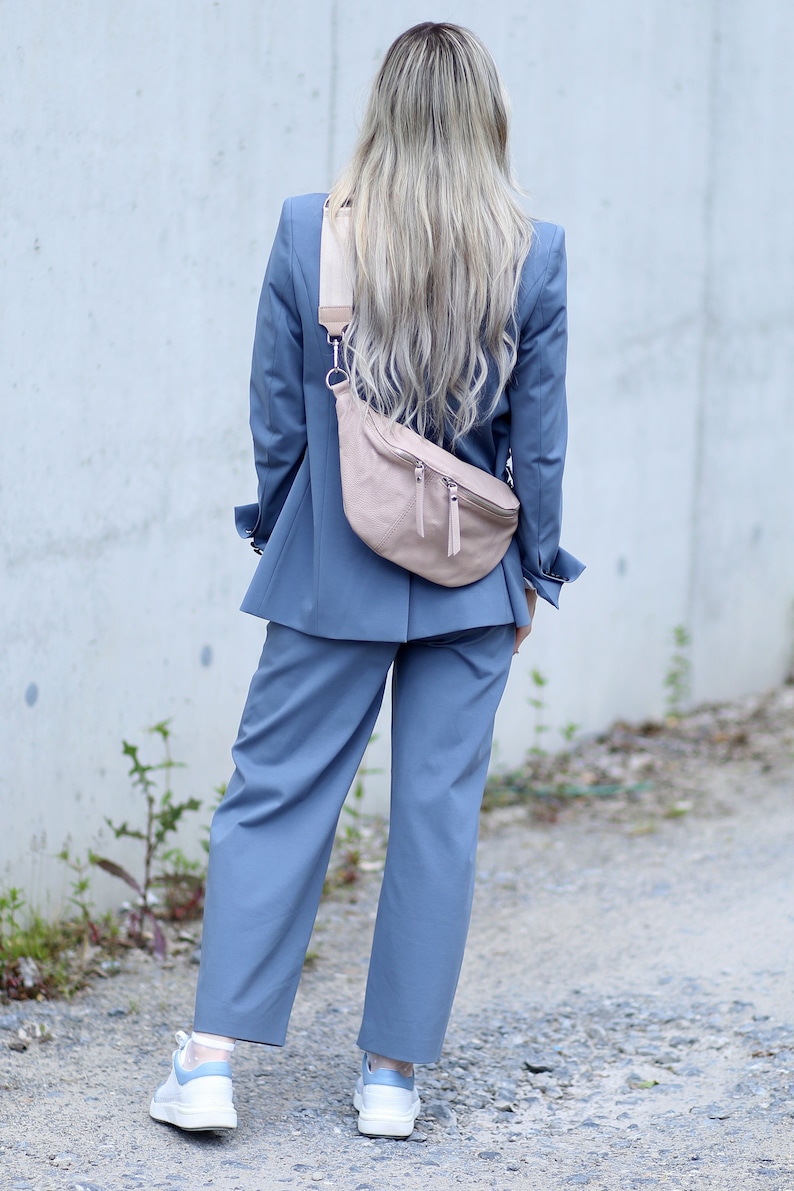 Op de afbeelding: Een lichtblauw pak bestaande uit een blazer en een broek met wijde pijpen, gecombineerd met een lichtroze heuptas. De heuptas heeft twee vakken met ritssluiting en een beige band. Witte sneakers met blauwe accenten maken de outfit compleet.
