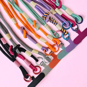 Può includere: Una collezione di cordini colorati per telefoni. I cordini presentano una varietà di colori, tra cui nero, rosa, arancione, viola e verde acqua. Ogni cordino ha un design annodato e un anello di metallo per il fissaggio a una custodia del telefono. Lo sfondo è rosa.