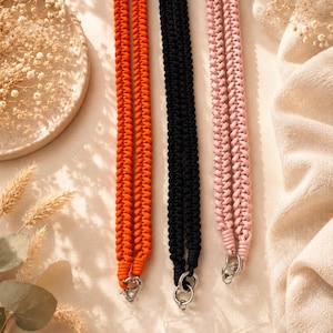 May include: Three long lanyards featuring a braided cord design in bright orange, solid black, and soft pink. Each lanyard is finished with a silver-toned metal ring and a clear, rectangular plastic attachment at the bottom for holding items.