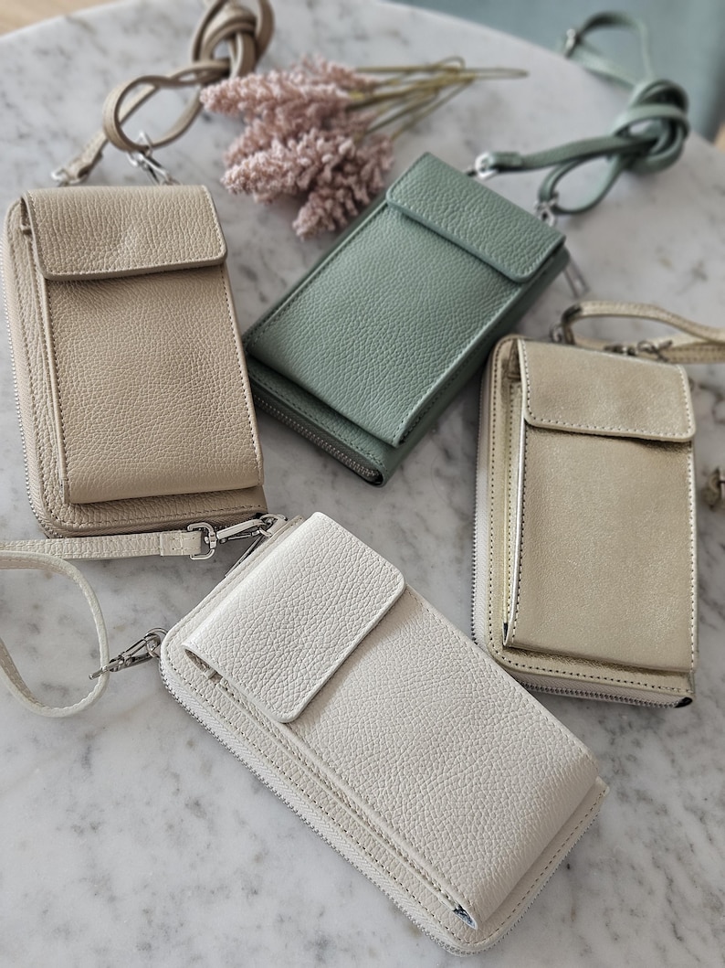 May include: Four leather crossbody phone bags in beige, sage green, off-white, and gold. Each bag has a flap closure, a zippered compartment, and a detachable strap. The bags are displayed on a marble surface.