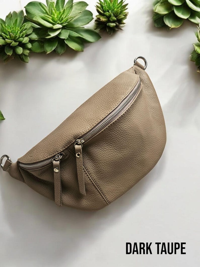 Op de afbeelding: Een donker taupe leren heuptas met twee compartimenten met ritssluiting en zilverkleurige hardware. De tas heeft een gebogen vorm en een getextureerd oppervlak. De tekst "DARK TAUPE" wordt onderaan de afbeelding weergegeven.