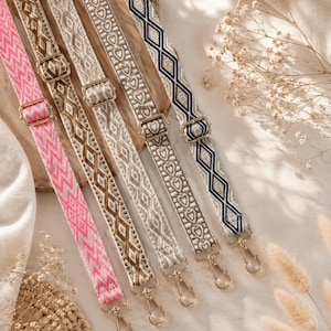 May include: Five adjustable bag straps featuring woven geometric and heart patterns in pink, brown, beige, and navy blue. Each strap includes gold-toned metal hardware, including sliding buckles for length adjustment and swivel lobster claw clasps.