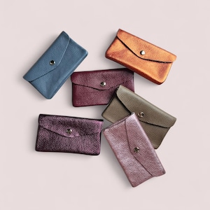 May include: Six rectangular envelope-style pouches with a pebbled texture and a single silver-tone snap button closure. The pouches are arranged in a cluster and feature metallic and matte finishes in blue, copper, burgundy, taupe, and pink tones.