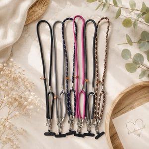 May include: Six phone lanyards featuring various cord styles, including solid black, solid gray, navy blue with white stripes, solid magenta, dark gray, and a brown and white diamond pattern, each with silver metal hardware and small leather accents.
