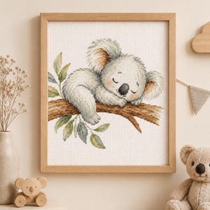 Puede incluir: Un bordado en punto de cruz enmarcado que muestra a un koala durmiendo sobre una rama de árbol marrón con hojas verdes. El koala tiene pelaje gris, orejas grandes y una nariz negra. La obra de arte está en un marco de madera clara.