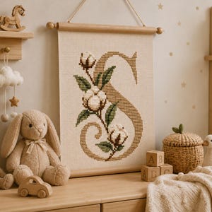 Op de afbeelding: Een rechthoekige wandhanger met een kruissteekontwerp van de letter S in een beige kleur, versierd met witte katoenbollen en groene bladeren. De stof is bevestigd aan een lichte houten stok met een ophangkoord aan de bovenzijde.