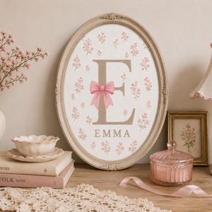Può includere: Un'opera d'arte da parete con cornice ovale che presenta una grande lettera E beige centrata su uno sfondo bianco con un motivo floreale rosa ripetuto. Un fiocco rosa è legato attorno alla lettera E e il nome EMMA è stampato in beige sotto.