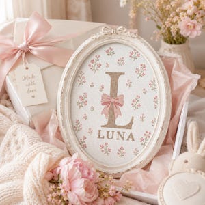 May include: An oval-shaped white frame containing a cross-stitch embroidery of the letter L and the name LUNA in brown, accented with a pink bow and small pink floral patterns on a white fabric background, presented in a gift box with ribbon.