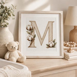 Op de afbeelding: Een vierkant ingelijst kruissteek kunstwerk met een grote, beige, getextureerde letter M, versierd met witte katoenbollen en groene bladeren. Het borduurwerk is geplaatst tegen een witte achtergrond in een licht houten lijst.