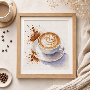 Może przedstawiać: Oprawiony haft krzyżykowy przedstawiający białą ceramiczną filiżankę z kawą z wzorem latte art na pasującym spodku. Wzór zawiera rozsypane brązowe ziarna kawy i zmieloną kawę na białym tle z delikatnym niebieskim cieniem pod spodem.