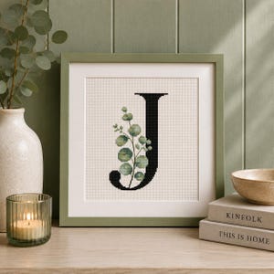 May include: A square framed cross-stitch artwork featuring a large black letter J decorated with a green eucalyptus branch. The embroidery is set against a light cream-colored fabric background and enclosed in a simple sage green wooden frame.