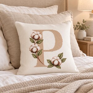 Peut inclure: Un coussin carré en tissu blanc texturé orné d'une grande lettre P de couleur beige brodée au point de croix. La lettre est décorée de trois grappes de fleurs de coton blanches accompagnées de feuilles vertes détaillées.
