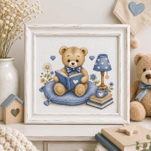 Peut inclure: Une broderie au point de croix encadrée représentant un ours en peluche brun clair assis sur un coussin bleu, lisant un livre bleu. L'ours porte un nœud papillon bleu à pois. À côté se trouvent une lampe bleue, des livres et une tasse bleue.
