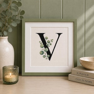 Op de afbeelding: Een vierkant ingelijst kruissteek kunstwerk met een grote zwarte hoofdletter V, versierd met een takje met groene eucalyptusbladeren. Het borduurwerk is geplaatst op een lichtcrèmekleurige stoffen achtergrond in een saliegroene lijst.