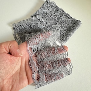 May include: A length of gray lace fabric with a delicate floral pattern. The lace is folded and held in a hand, showcasing its intricate design and texture. The fabric has a scalloped edge.