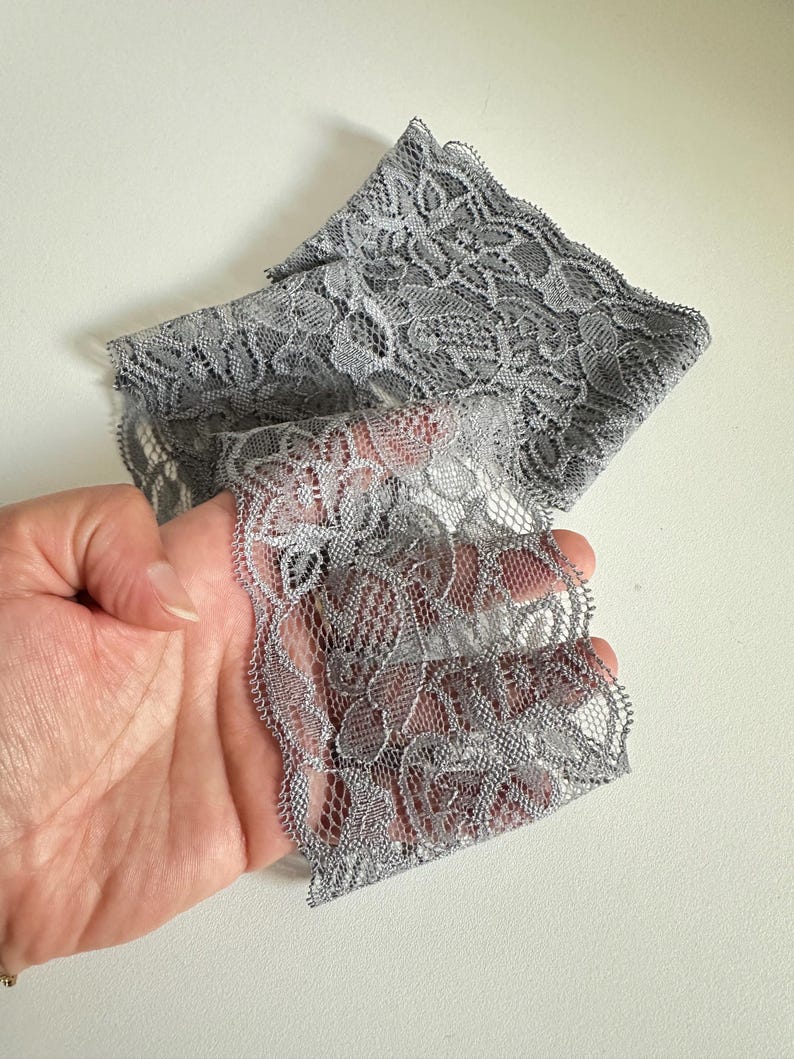 May include: A length of gray lace fabric with a delicate floral pattern. The lace is folded and held in a hand, showcasing its intricate design and texture. The fabric has a scalloped edge.