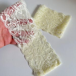 May include: Two pieces of delicate lace trim in off-white. The lace features a floral pattern with scalloped edges. One piece is a long strip, and the other is a shorter, folded piece, both on a white background.