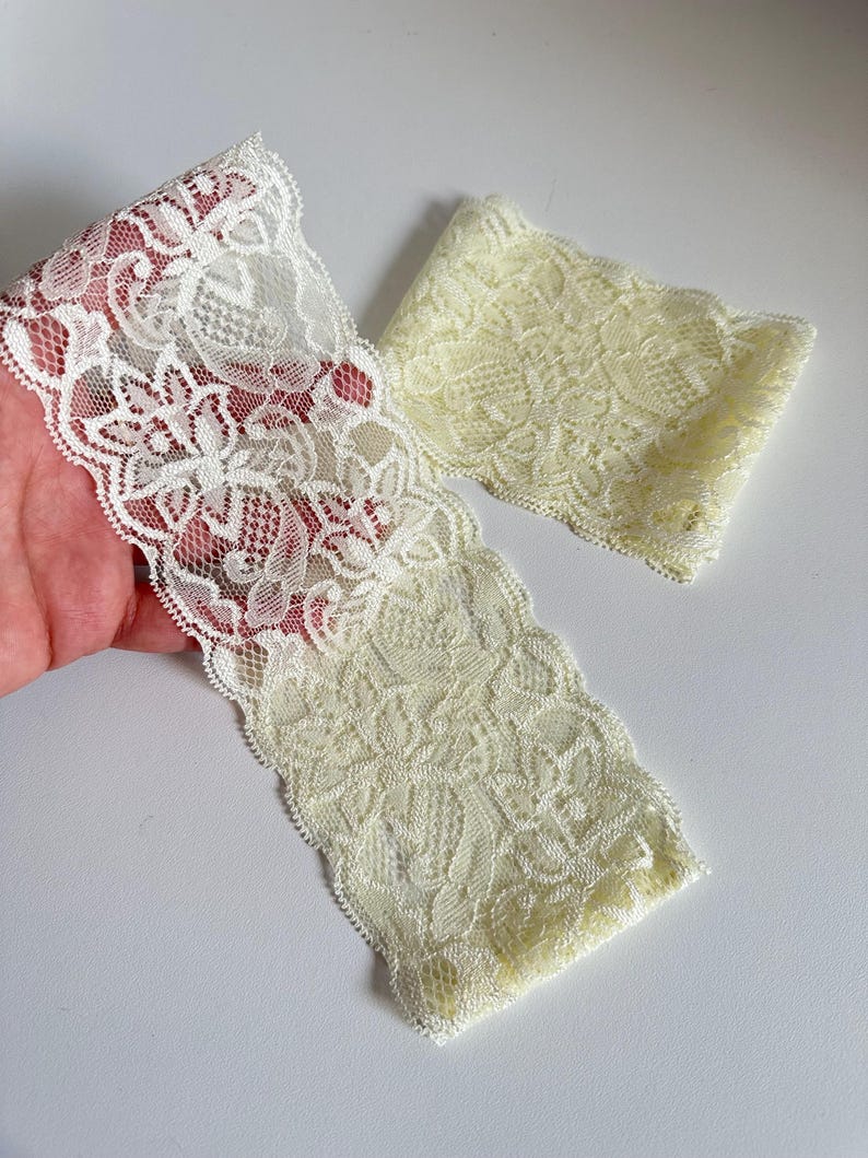 May include: Two pieces of delicate lace trim in off-white. The lace features a floral pattern with scalloped edges. One piece is a long strip, and the other is a shorter, folded piece, both on a white background.