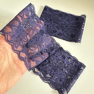 May include: Close-up of three pieces of dark purple lace fabric. The lace has a floral pattern and a scalloped edge. The fabric is held in a hand against a white background.