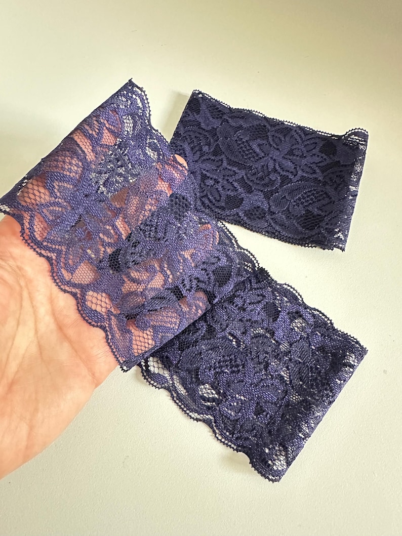 May include: Close-up of three pieces of dark purple lace fabric. The lace has a floral pattern and a scalloped edge. The fabric is held in a hand against a white background.