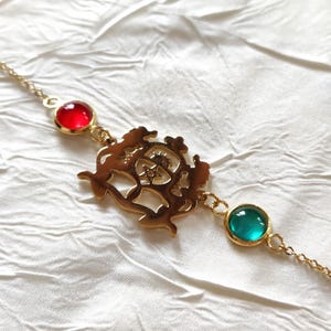 Op de afbeelding: Gouden ketting met een gedetailleerde hanger met een gestileerd ontwerp en twee edelstenen, een rode en een teal.