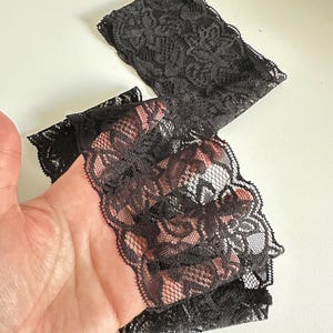 May include: Black lace fabric with a floral pattern. The delicate material is folded and appears to be a trim or edging for clothing or crafts. The lace has a scalloped edge.