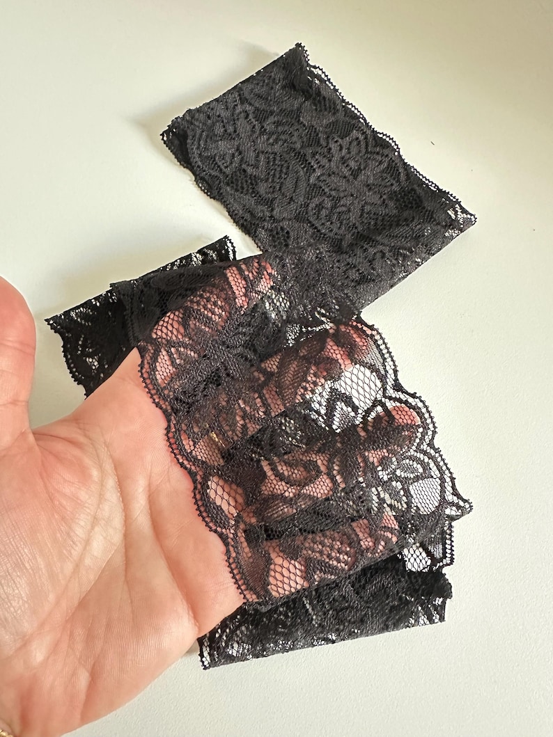 May include: Black lace fabric with a floral pattern. The delicate material is folded and appears to be a trim or edging for clothing or crafts. The lace has a scalloped edge.
