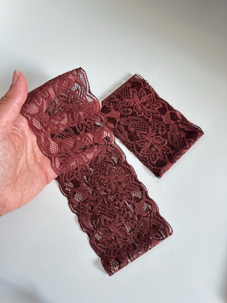 May include: Two pieces of burgundy lace fabric with a floral pattern. One piece is folded, and the other is rectangular. The lace has a scalloped edge and is displayed on a white background.